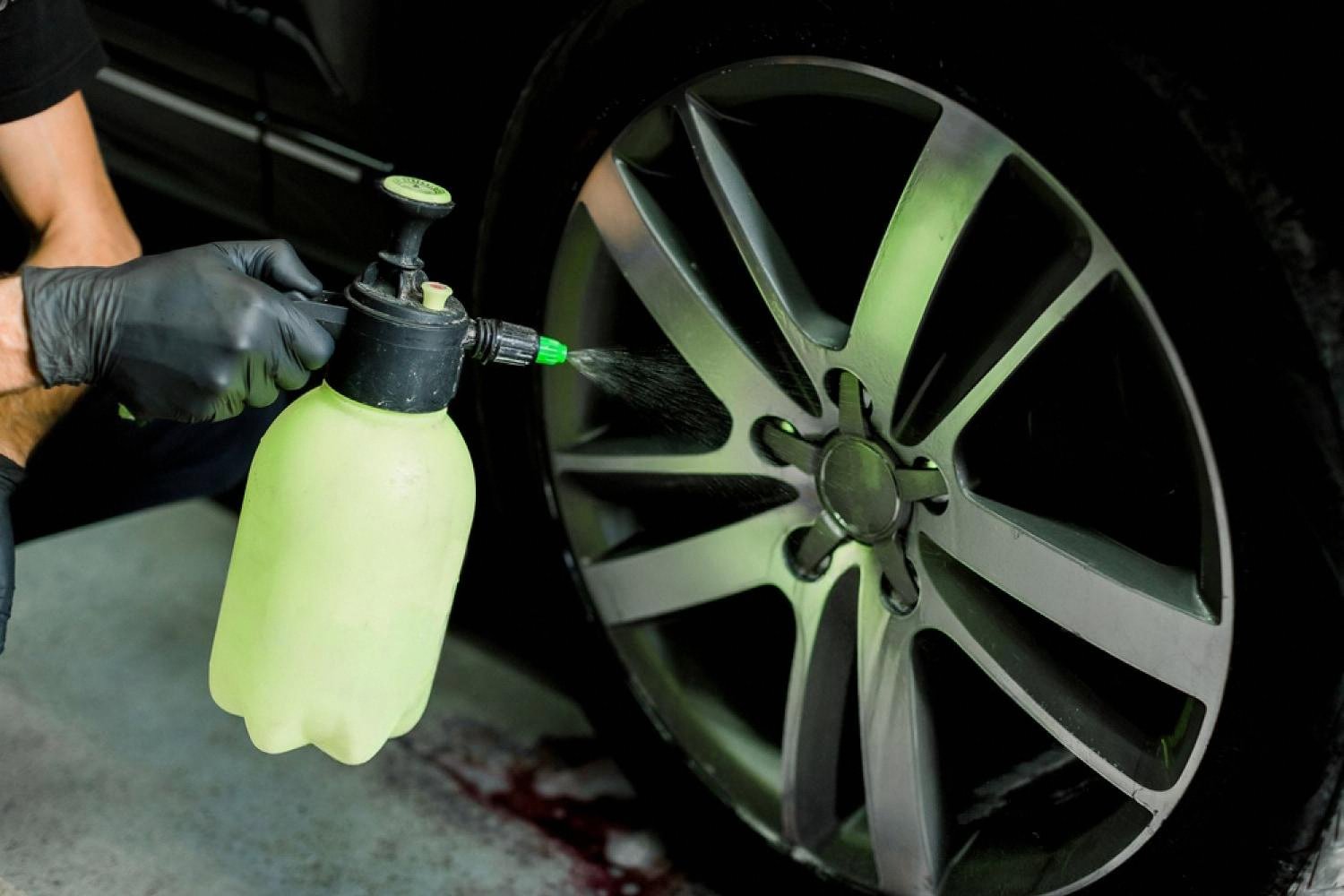 Gloved hand spraying green wheel cleaner onto a black alloy car rim